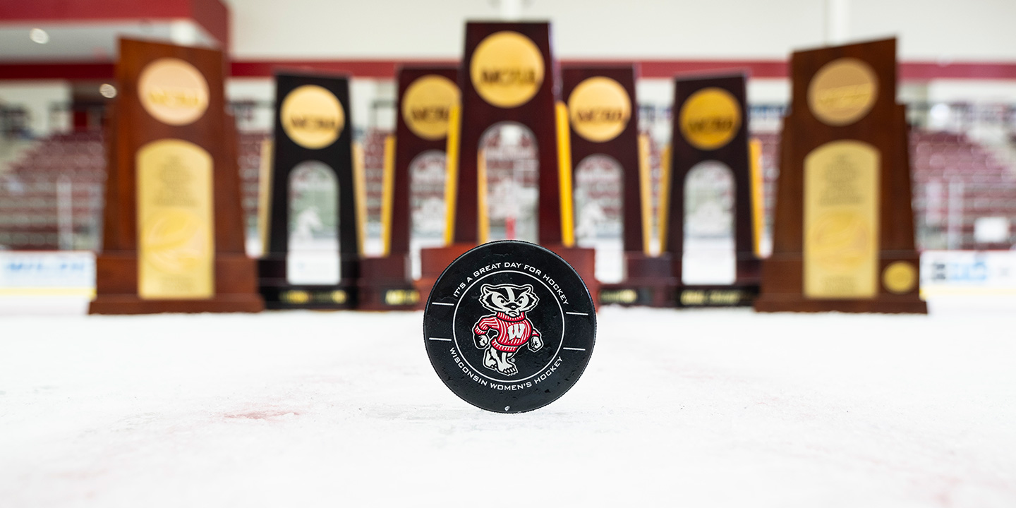 A hockey puck with Bucky Badger is displayed in front of seven hockey trophies.