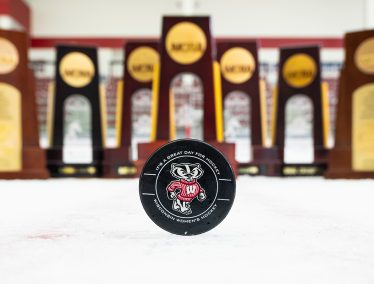 A hockey puck with Bucky Badger is displayed in front of seven hockey trophies.
