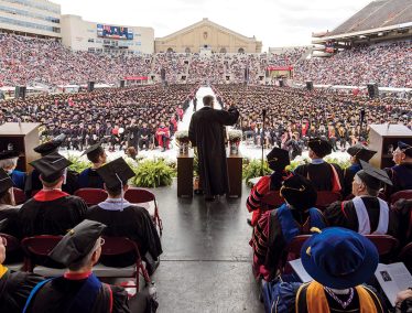 The view from the back of the stage at commencement at Camp Randall; the commencement speaker gesturing to hundreds of seated graduates in front of him.