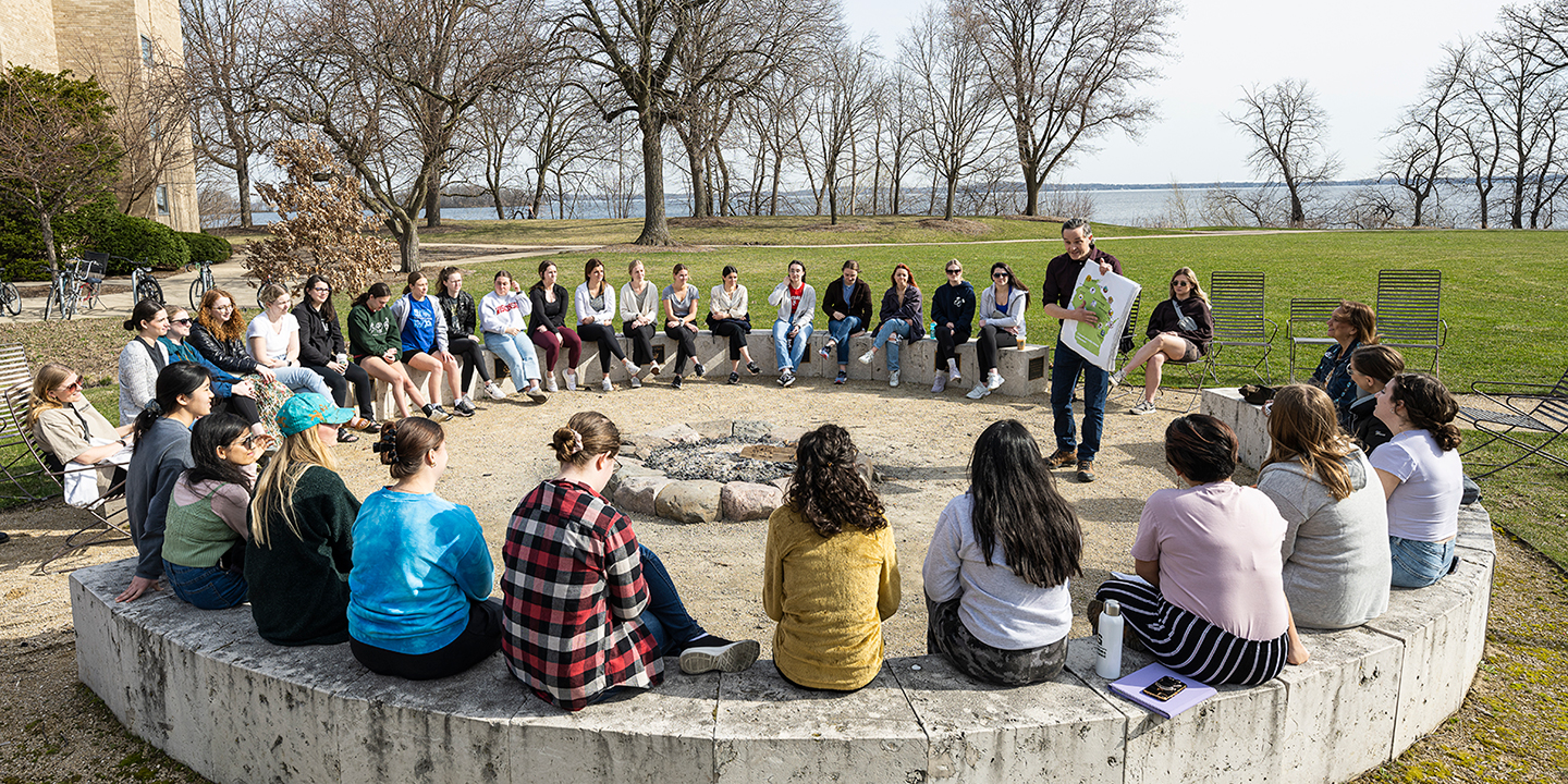 A First Nations Cultural Tour guide holds up a map of Wisconsin to a group sitting outdoors, in a circle.