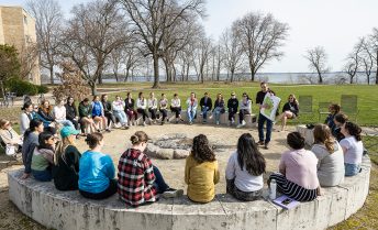 A First Nations Cultural Tour guide holds up a map of Wisconsin to a group sitting outdoors, in a circle.