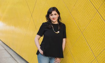 Marcela Guerrero, in a black shirt and jeans, leans against a yellow tiled wall.