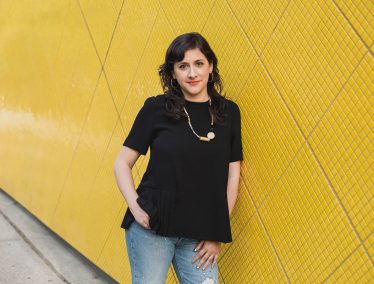 Marcela Guerrero, in a black shirt and jeans, leans against a yellow tiled wall.