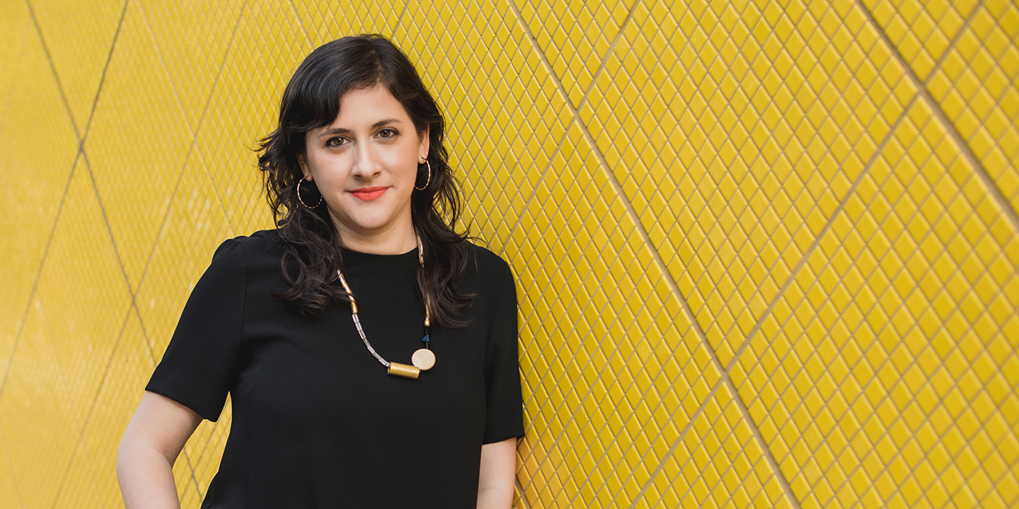 Marcela Guerrero, in black, leans against a yellow tiled wall.