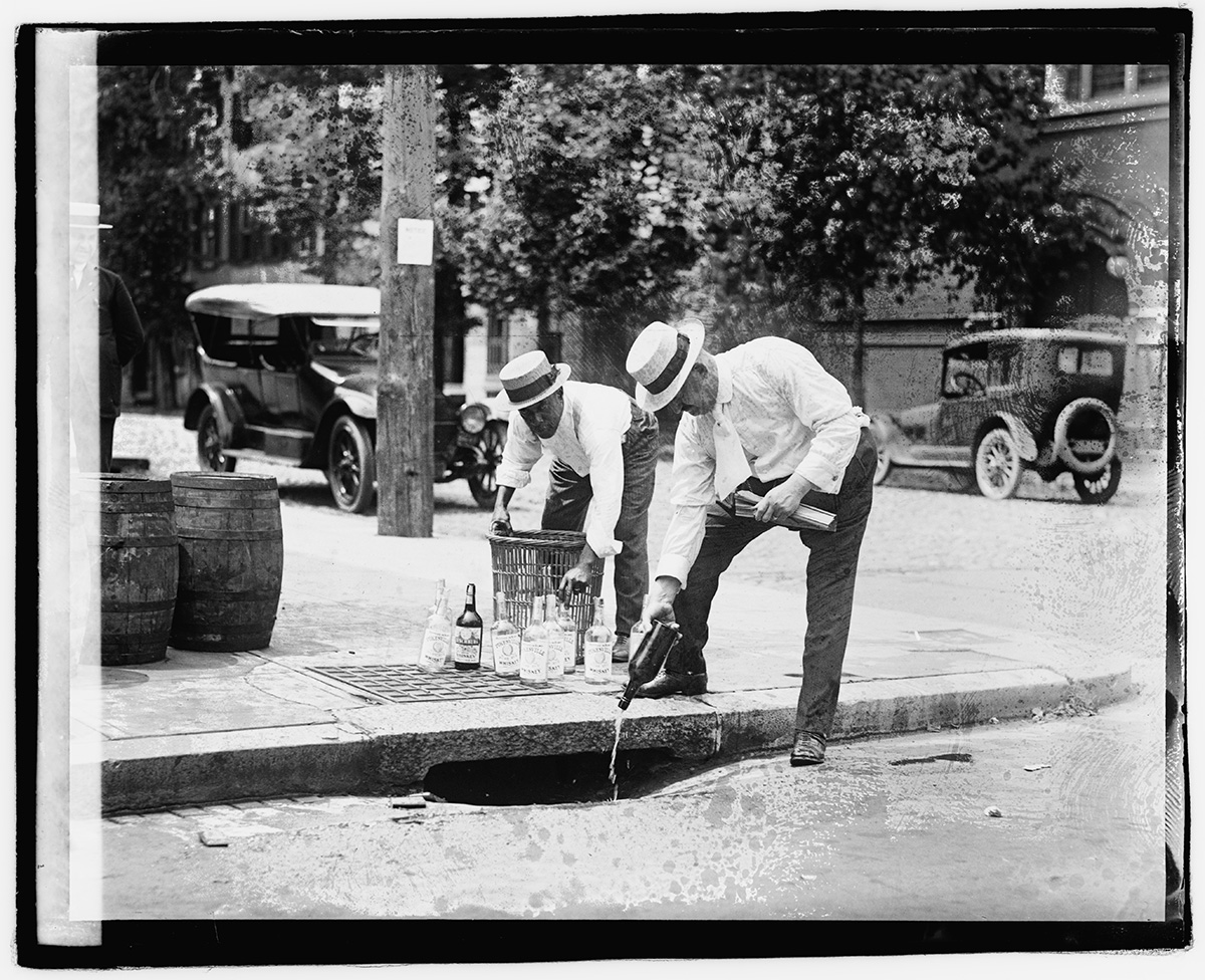 Black and white photograph of two men pouring whiskey down a street gutter.