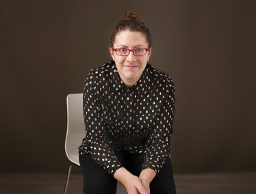 Rachel Willson-Broyles, dressed in dark colors, sitting on a white chair in a dark room, leaning toward the camera.