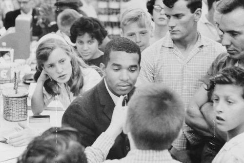 Black and white photo of a young Dion Diamond being accosted by a mob of white people at a 1960s drugstore counter.