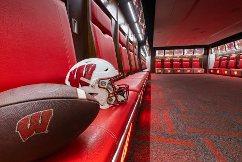 A football and Wisconsin Badger football helmet sit on a red bench in the football locker room.