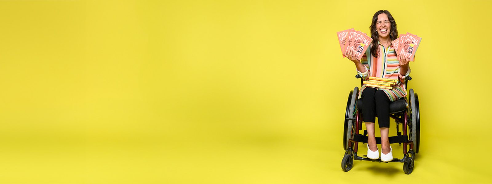 An image of a woman against a bright yellow background. She is sitting in a wheelchair and holding several copies of Taussig's book.