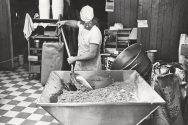 A man stirs a giant mixture of cookie batter in a black and white photo.