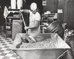 A man stirs a giant mixture of cookie batter in a black and white photo.