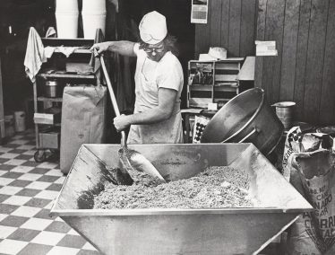 A man stirs a giant mixture of cookie batter in a black and white photo.