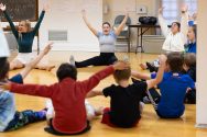 A dance instructor leads a group of children stretching their arms and legs on the floor