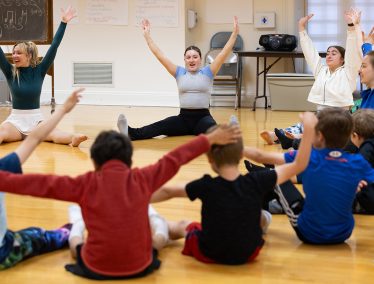 A dance instructor leads a group of children stretching their arms and legs on the floor