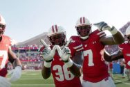 Badger football players smile and flash the 'W' sign on the sidelines of a game at Camp Randall