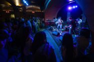 A band performs on stage under blue and purple lights in front of an audience at a dimly lit venue with arched ceilings.