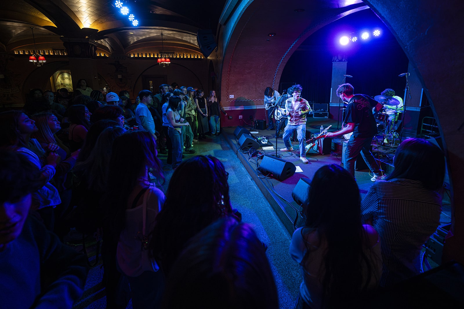 A band performs on stage under blue and purple lights in front of an audience at a dimly lit venue with arched ceilings.