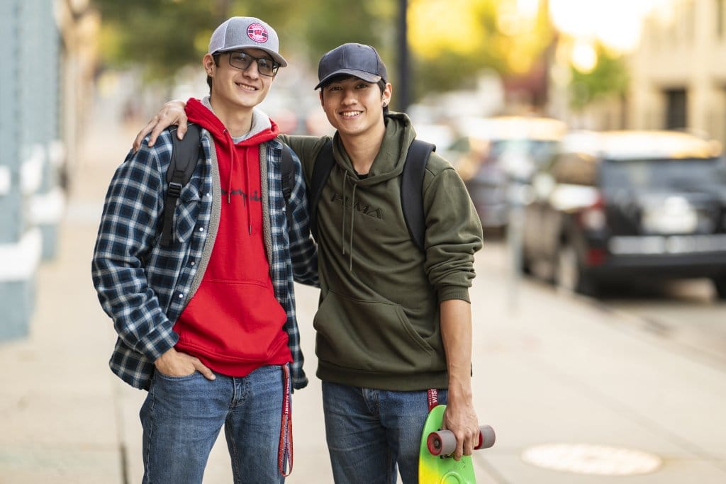 Two young men stand side by side, wearing baseball hats and informal clothes. One carries a skateboard.