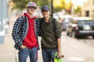 Two young men stand side by side, wearing baseball hats and informal clothes. One carries a skateboard.