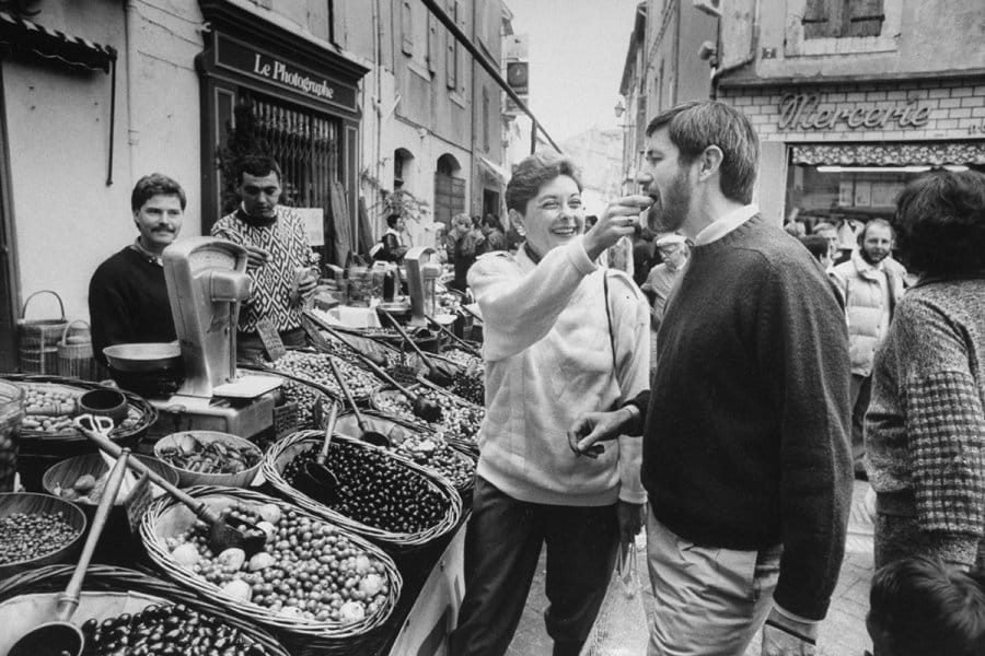 Black and white photo of Patricia and Walter Wells sampling olives at a street market in France