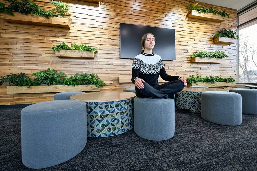 A woman sits cross-legged with her eyes closed on a round, gray ottoman in a modern lounge area with several planters mounted on the wall behind her.