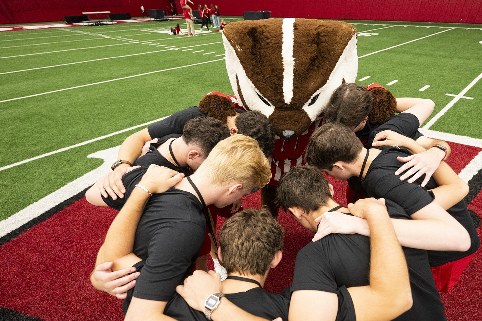 A group of students in black shirts huddled together on a football field with Bucky Badger in his classic red and white striped shirt.