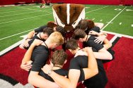 A group of students in black shirts huddled together on a football field with Bucky Badger in his classic red and white striped shirt.