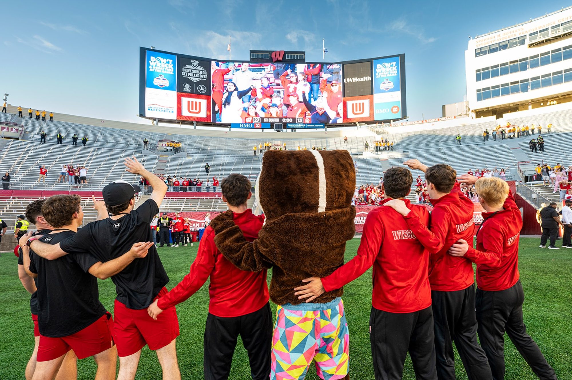A group of Buckys -- but only one in the mascot suit -- stand arm-in-arm on a football field facing the large stadium screen at Camp Randall with their backs to the camera.