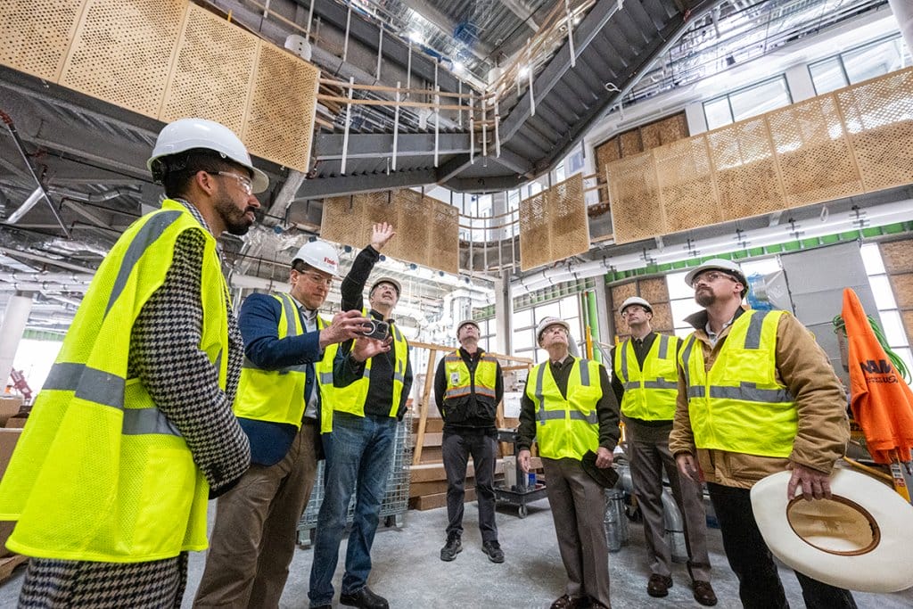 A group of people wearing hard hats and high-visibility vests are gathered inside a large, modern construction site, with one person gesturing upwards towards the ceiling.