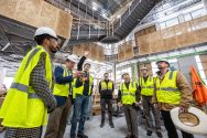 A group of people wearing hard hats and high-visibility vests are gathered inside a large, modern construction site, with one person gesturing upwards towards the ceiling.