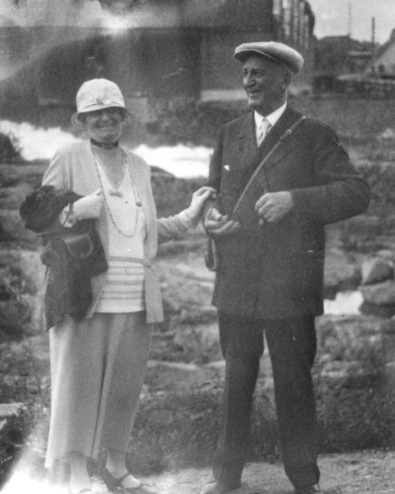 A black and white photo of Annie in a light-colored dress and hat holding a handbag, and Magnus wearing a dark suit with a cap and a bag over one shoulder.