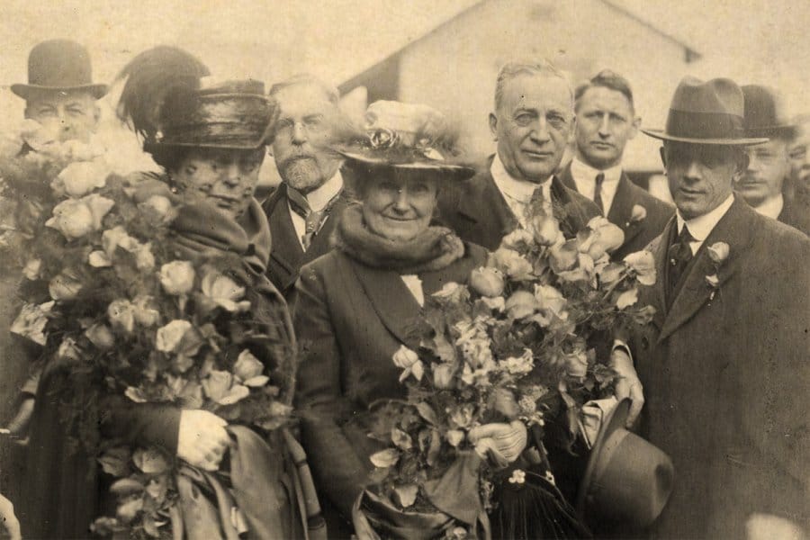 Sepia-toned photograph of a small group of people in early 20th-century attire, the two women holding large bouquets of flowers.