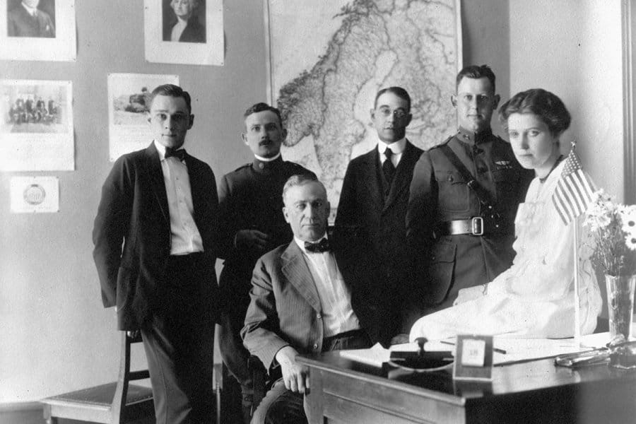 Black and white photograph of six people in a room with a large map of Scandinavia on the wall and an American flag on the desk.