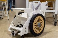 A small, custom-made wheelchair made from wood painted white and containing a harness, foot straps and extra big wheels.