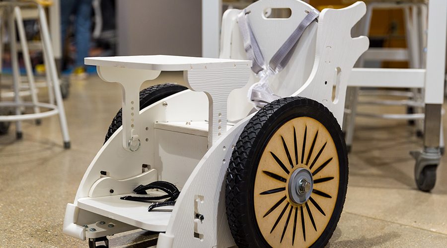 A small, custom-made wheelchair made from wood painted white and containing a harness, foot straps and extra big wheels.