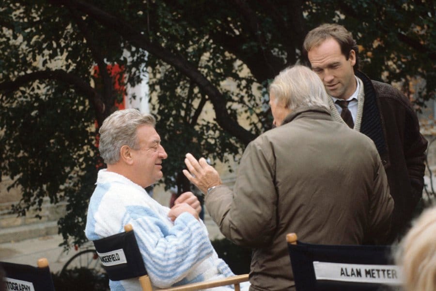 Rodney Dangerfield wearing a bathrobe and sitting in a director's chair with his name on it, discussing the film with two men in winter coats.