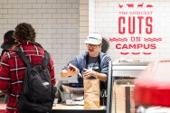 A person checks out at a deli counter, where another person is handing over a brown paper bag; the wall behind them has the text "The choicest cuts on campus" in red letters.