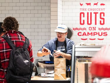 A person checks out at a deli counter, where another person is handing over a brown paper bag; the wall behind them has the text "The choicest cuts on campus" in red letters.