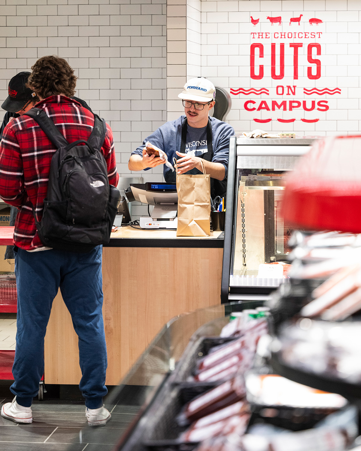 A person checks out at a deli counter, where another person is handing over a brown paper bag; the wall behind them has the text "The choicest cuts on campus" in red letters.