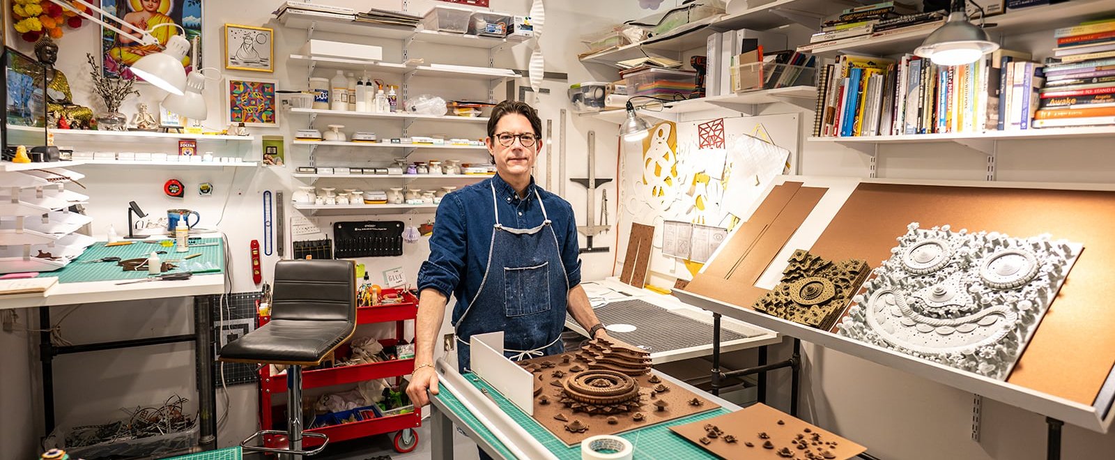 Michael Velliquette stands in a well-organized workshop filled with various art supplies, tools, and books, with intricate artwork displayed on the tables and walls.