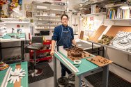 Michael Velliquette stands in a well-organized workshop filled with various art supplies, tools, and books, with intricate artwork displayed on the tables and walls.