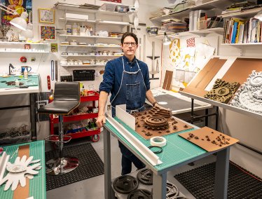 Michael Velliquette stands in a well-organized workshop filled with various art supplies, tools, and books, with intricate artwork displayed on the tables and walls.