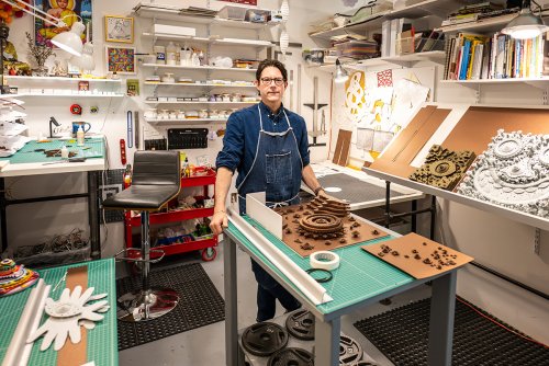 Michael Velliquette stands in a well-organized workshop filled with various art supplies, tools, and books, with intricate artwork displayed on the tables and walls.