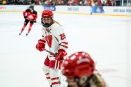 Hockey player in a white and red uniform with number 26 and a captain's 'C' on the chest skating on the ice, with another player in red and black visible in the background and a teammate visible in the foreground.