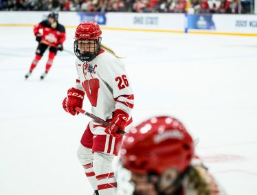 Hockey player in a white and red uniform with number 26 and a captain's 'C' on the chest skating on the ice, with another player in red and black visible in the background and a teammate visible in the foreground.