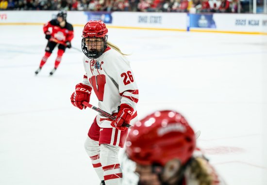 Hockey player in a white and red uniform with number 26 and a captain's 'C' on the chest skating on the ice, with another player in red and black visible in the background and a teammate visible in the foreground.