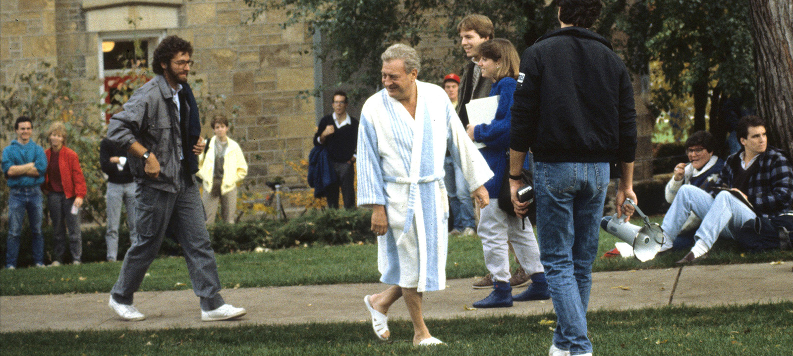 A group of people gathered outdoors on Bascon Hill, with Rodney Dangerfield (as character Thornton Melon) in a bathrobe and slippers among casually dressed students and actors.