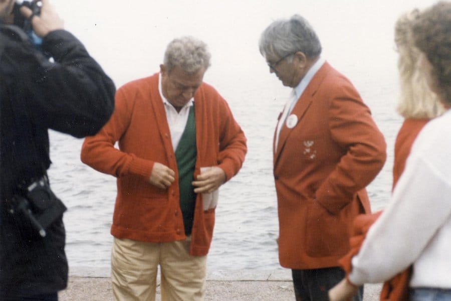 Two men stand near Lake Mendota. Rodney Dangerfield wears a red UW letter sweater and the other man wears a red jacket with a UW pin on his lapel and Bucky Badger on the pocket.