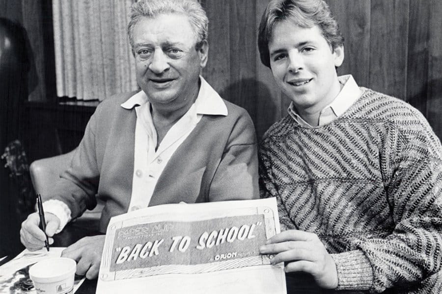 Dangerfield and another person sitting side by side, who holds a sign that reads "BACK TO SCHOOL" from Orion Pictures.
