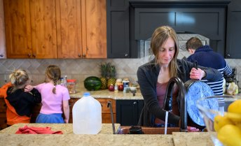 A kitchen scene with Jessica Calarco washing a large pot at the sink, two children standing at the counter, and her husband cooking at the stove, surrounded by items like a gallon of milk, pineapples, watermelon, and kitchen utensils.